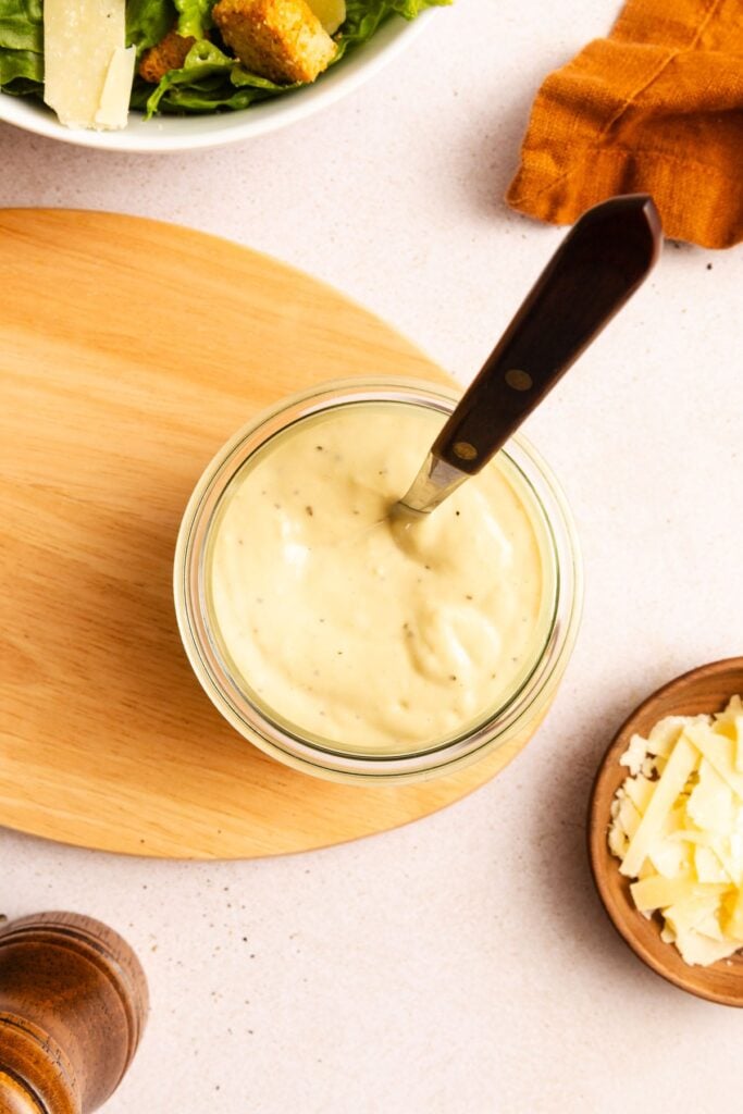 A jar of homemade Caesar salad dressing with a spoon in the bowl, placed on a wooden board beside a bowl of salad and a small dish of shaved Parmesan cheese.