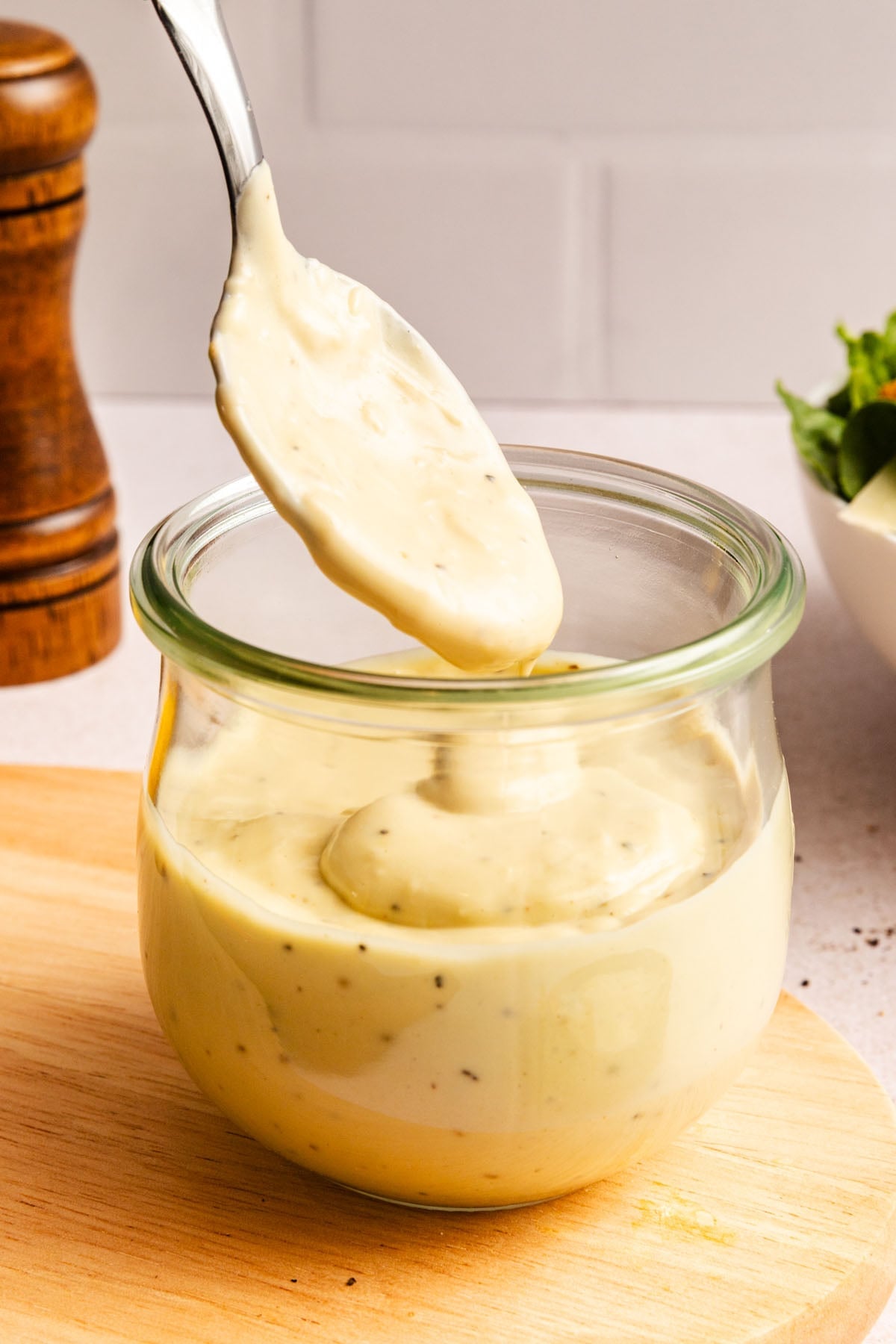 A spoon lifts creamy, herb-flecked dressing from a glass jar, placed on a wooden board with a pepper mill and a bowl of greens in the background.