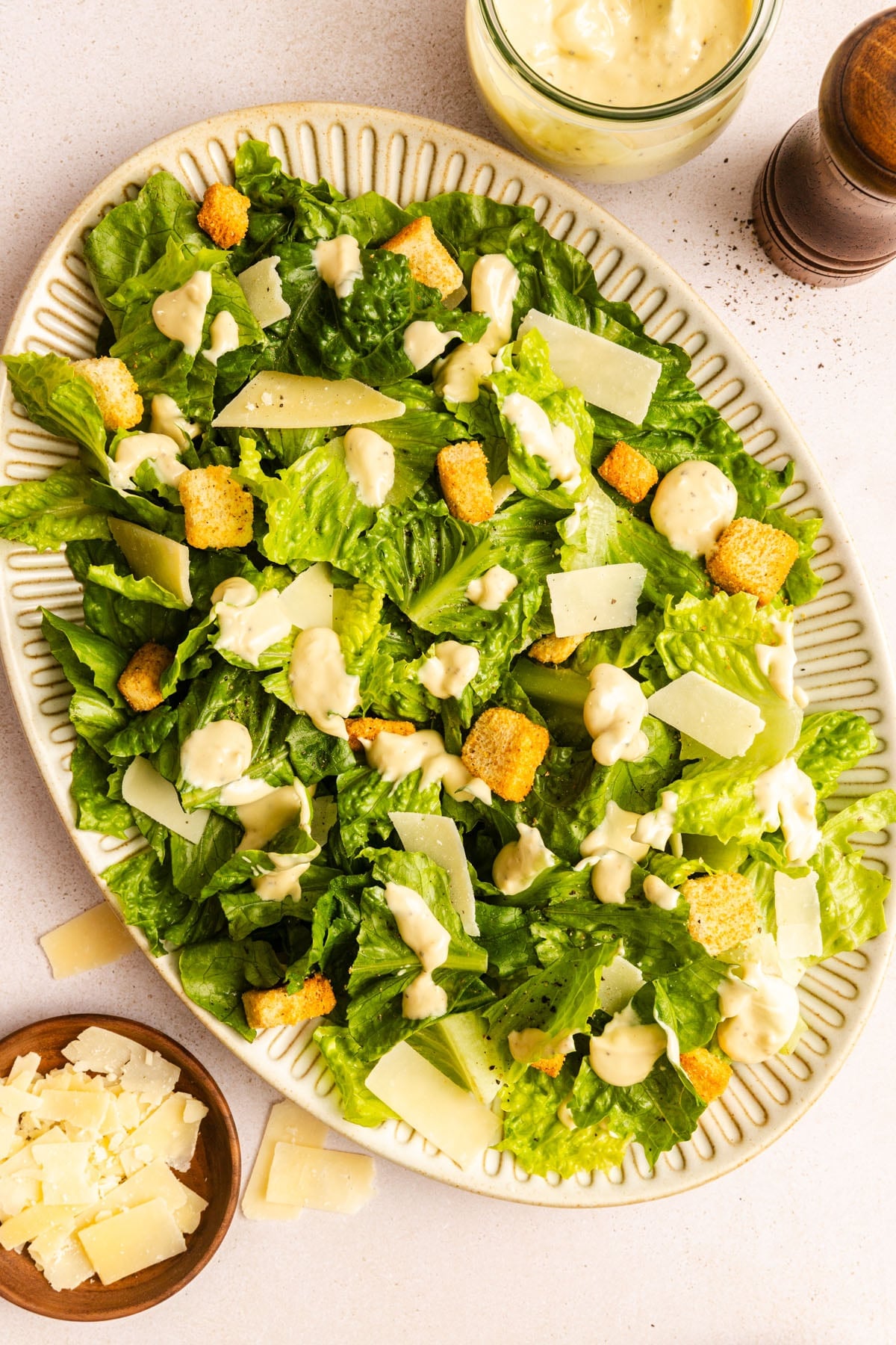 A plate of Caesar salad with romaine lettuce, croutons, shaved Parmesan cheese, and creamy Caesar dressing. A small bowl of cheese and a jar of dressing are nearby.