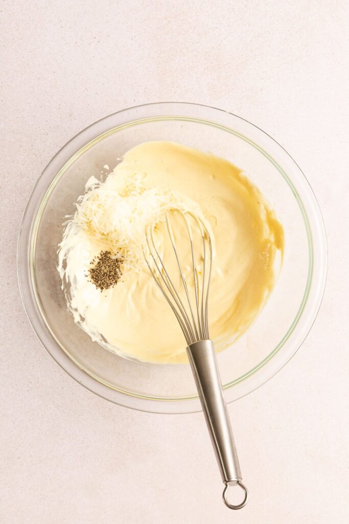 Whisking black pepper into creamy salad dressing in a glass bowl, viewed from above.