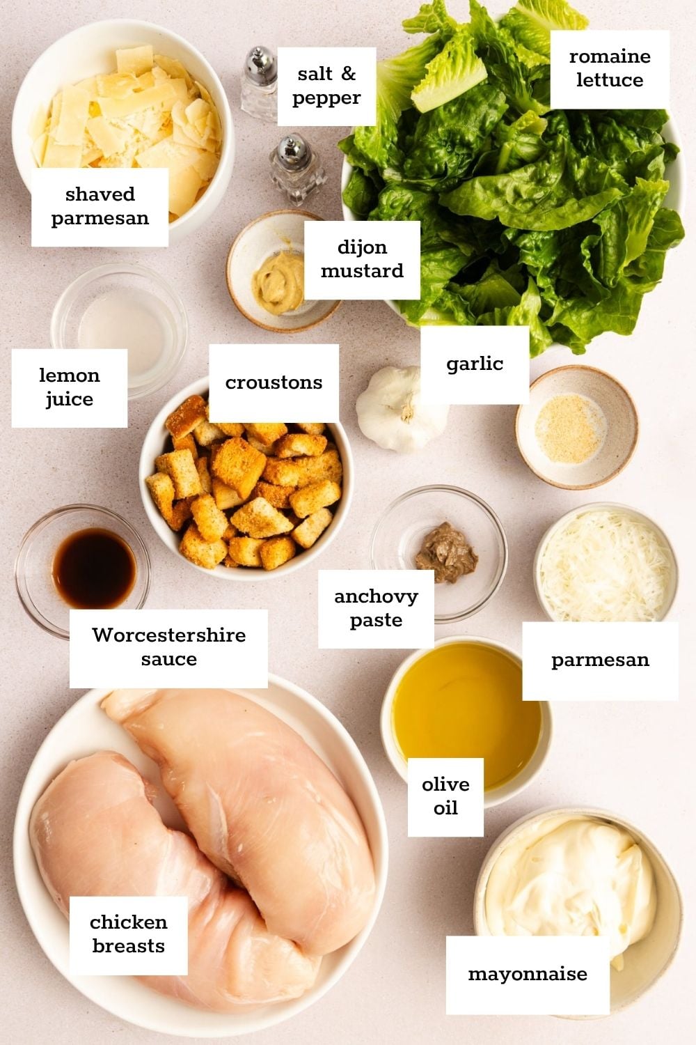 Ingredients for chicken Caesar salad are arranged on a countertop, including chicken breasts, romaine lettuce, croutons, parmesan, mayonnaise, olive oil, and various seasonings.