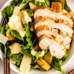 A bowl of Caesar salad with grilled, sliced chicken breast, romaine lettuce, croutons, shaved parmesan cheese, and Caesar dressing. A fork rests on the side of the bowl.