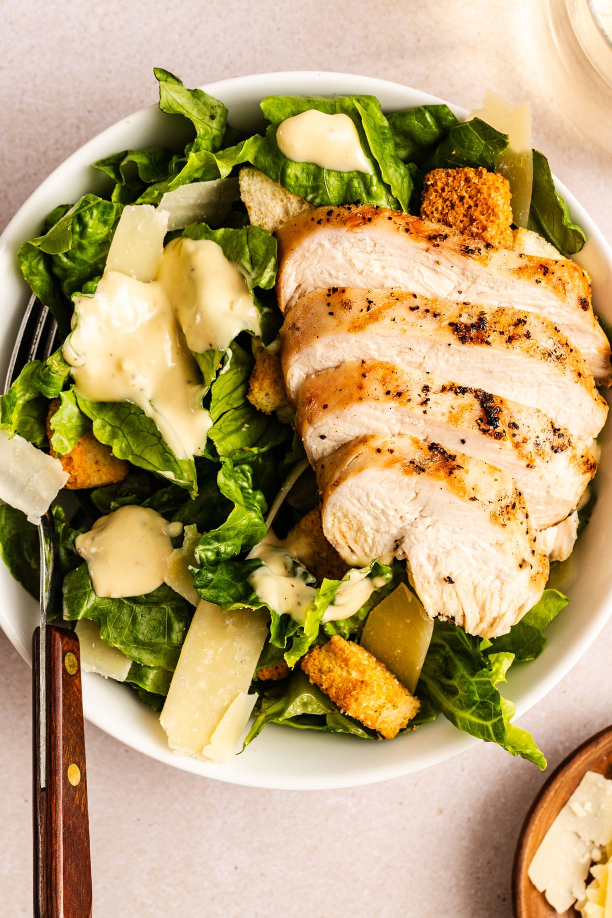 A bowl of Caesar salad with grilled, sliced chicken breast, romaine lettuce, croutons, shaved parmesan cheese, and Caesar dressing. A fork rests on the side of the bowl.