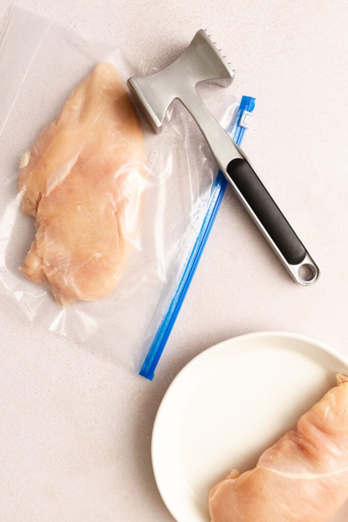 Raw chicken breast in a plastic bag next to a metal meat tenderizer and a plate with another piece of raw chicken breast on a light surface.