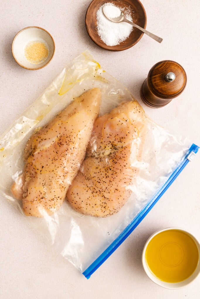 Two raw, seasoned chicken breasts in a resealable plastic bag, surrounded by bowls of salt, garlic powder, olive oil, and a pepper grinder on a light surface.