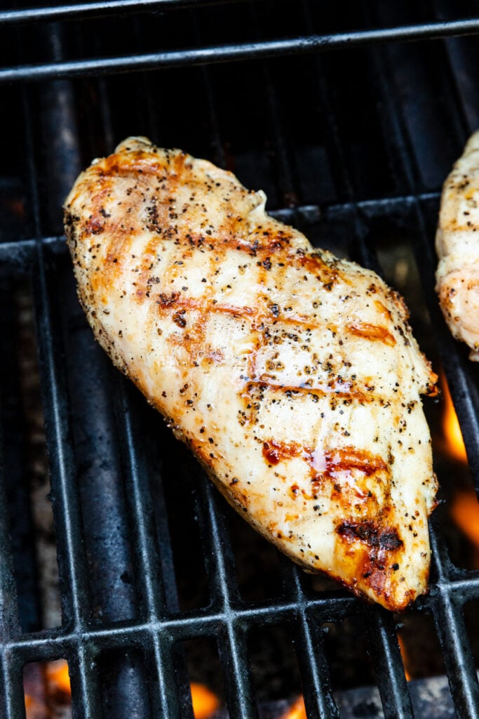 A seasoned chicken breast is cooking on a grill, showing grill marks and some flames visible below the grates.