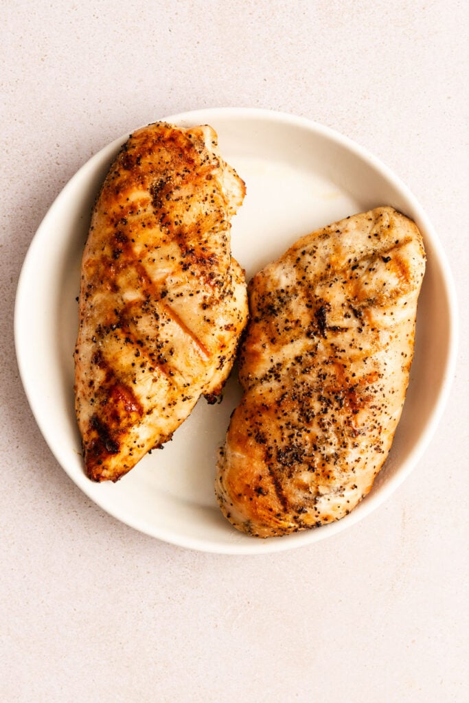 Two cooked, seasoned chicken breasts on a white plate, photographed from above against a light-colored background.