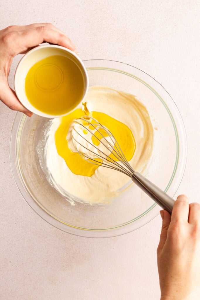 A person pours oil from a small bowl into a glass mixing bowl with batter while holding a whisk.