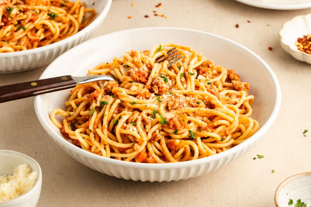 A white bowl filled with spaghetti topped with turkey Bolognese sauce, grated cheese, and fresh herbs, with a fork resting inside the bowl.
