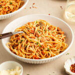 A bowl of spaghetti with tomato meat sauce is served on a table alongside grated cheese, chopped herbs, and a glass of white wine.