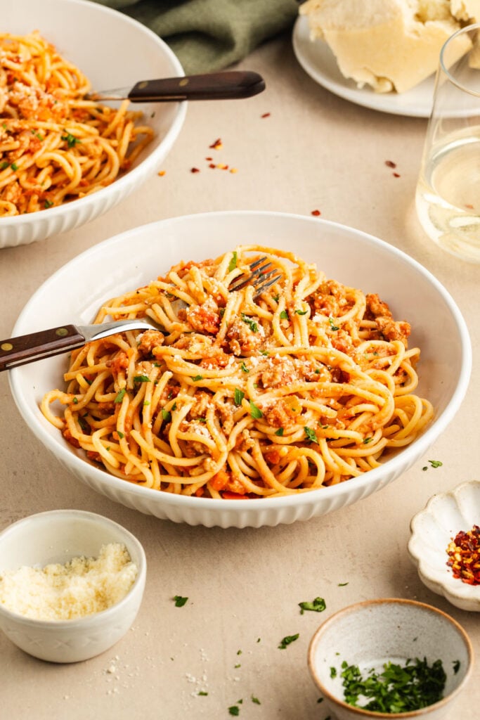 A bowl of spaghetti with tomato meat sauce is served on a table alongside grated cheese, chopped herbs, and a glass of white wine.