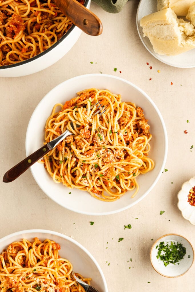 A bowl of spaghetti with meat sauce on a table, garnished with parsley, surrounded by more bowls of pasta, grated cheese, and bread.