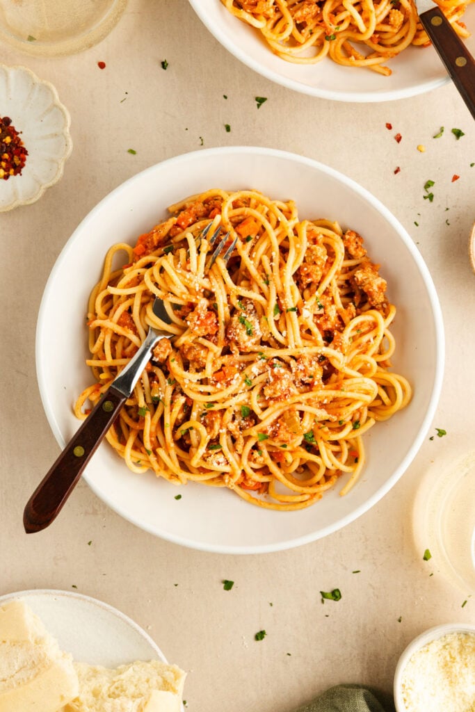 A white bowl of spaghetti with meat sauce, topped with grated cheese and parsley, sits on a beige table with a fork, surrounded by small dishes and pieces of bread.