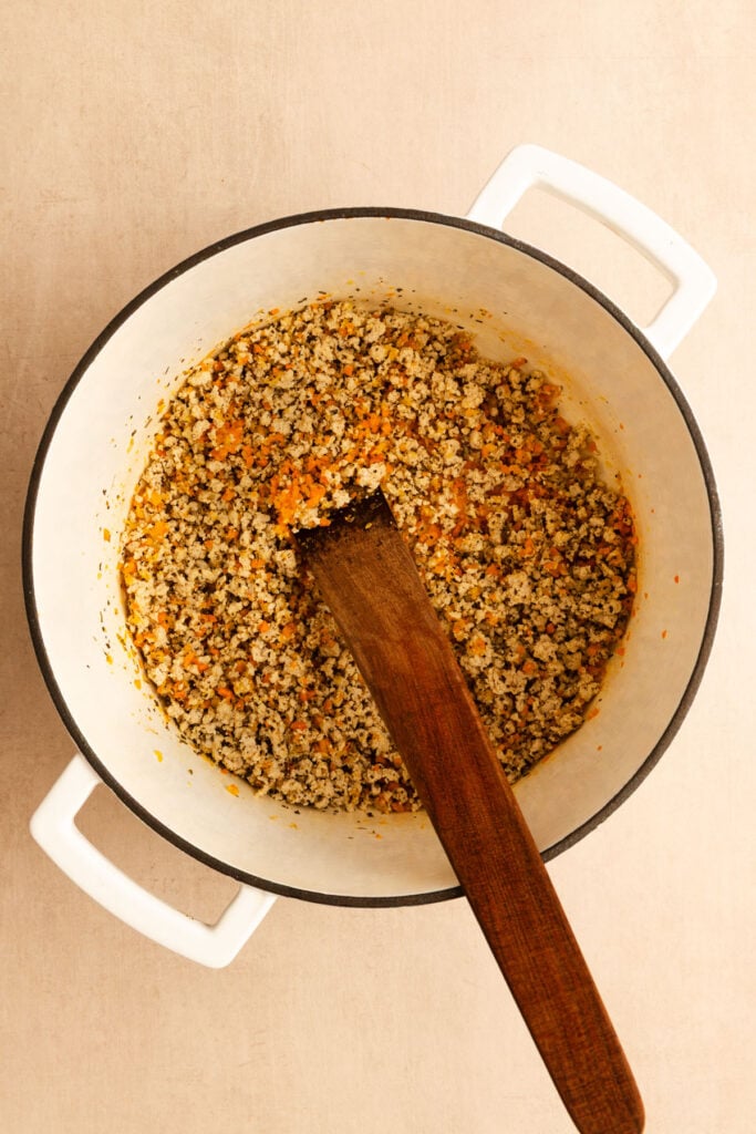 A white pot filled with cooked ground turkey, carrots, and herbs, with a wooden spatula resting inside.