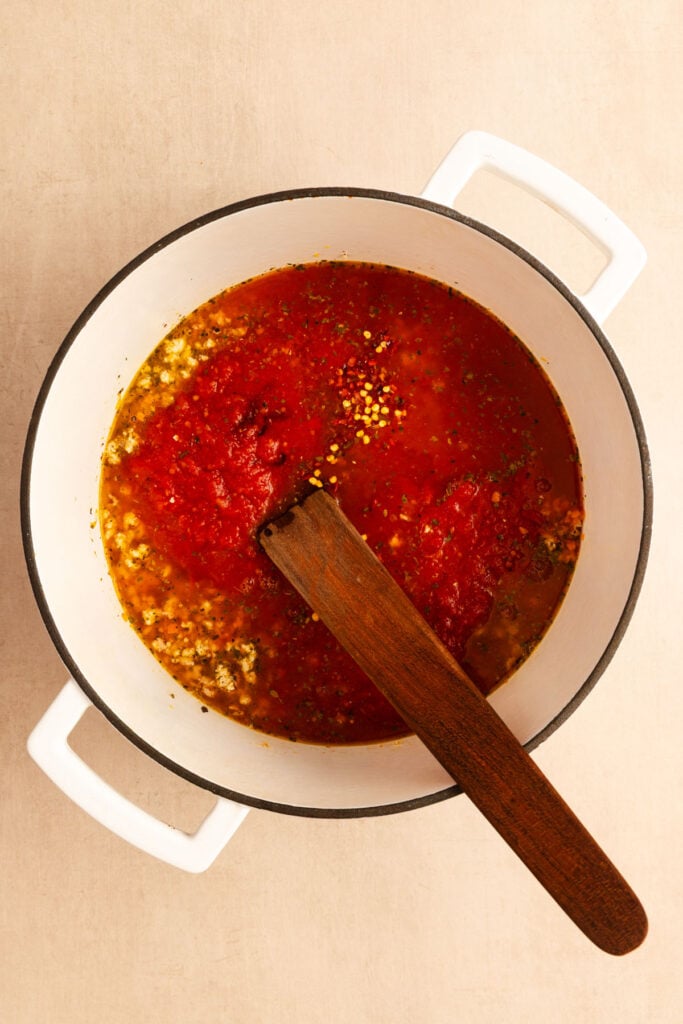 Overhead view of a wooden spoon in a large white Dutch oven that's filled with tomato sauce and fresh herbs.