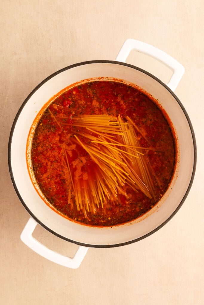 A white pot filled with tomato-based spaghetti sauce and uncooked spaghetti noodles, viewed from overhead.