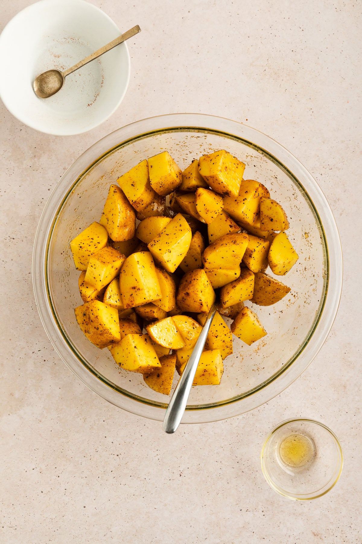 A glass bowl filled with seasoned, chopped yellow potatoes and a spoon, next to a small dish with seasoning residue and a small bowl of oil on a light countertop.