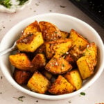 A white bowl filled with chunks of seasoned, roasted Yukon Gold potatoes, garnished with herbs, with a fork resting inside.