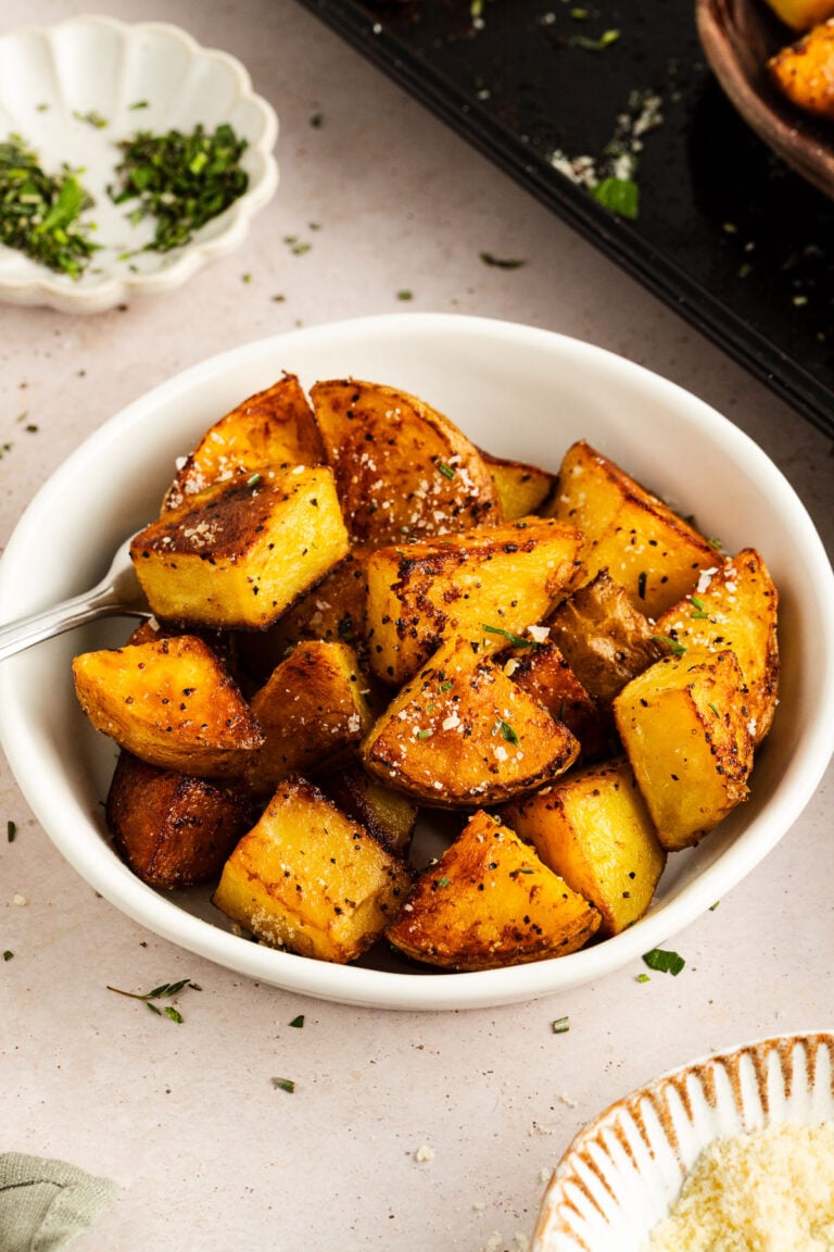 A white bowl filled with chunks of seasoned, roasted Yukon Gold potatoes, garnished with herbs, with a fork resting inside.