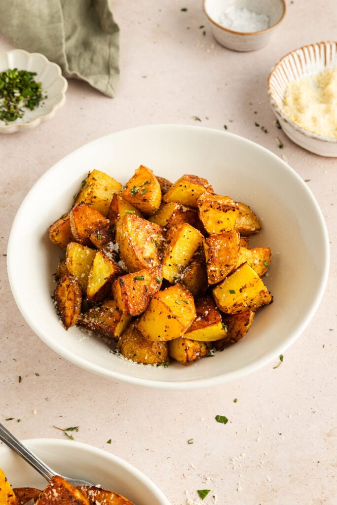 A white bowl filled with roasted Yukon Gold potato cubes topped with grated cheese and herbs, with small dishes of salt, cheese, and chopped herbs nearby.