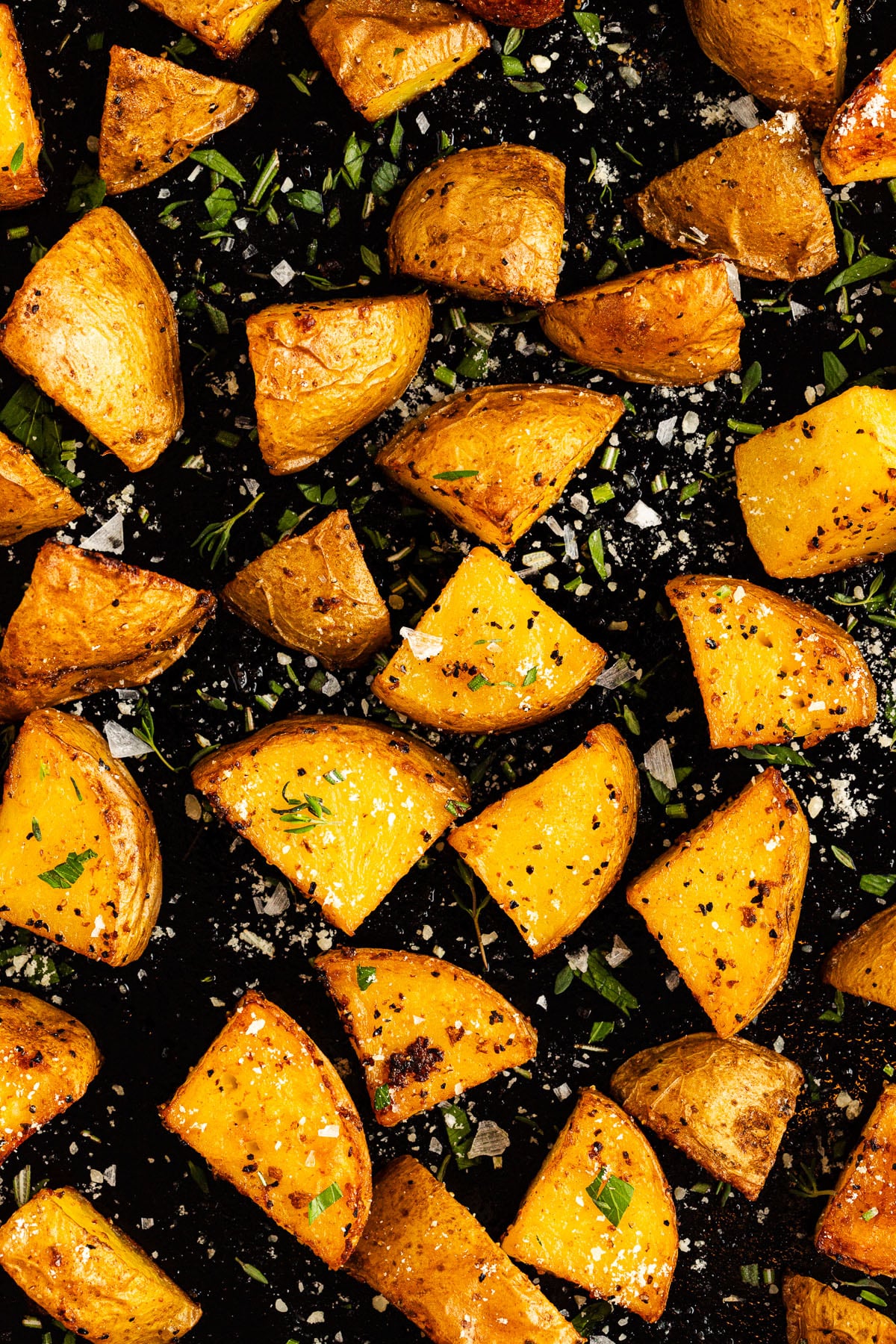 Golden roasted Yukon Gold potato wedges seasoned with herbs, black pepper, and coarse salt, arranged on a dark baking tray.
