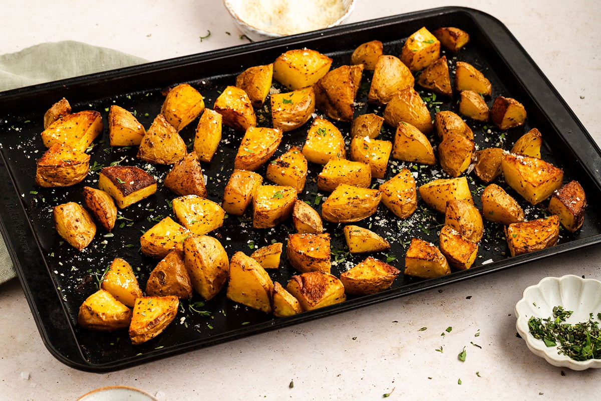 A baking tray filled with seasoned, oven roasted potato chunks, garnished with herbs and grated cheese, sits on a light-colored countertop.