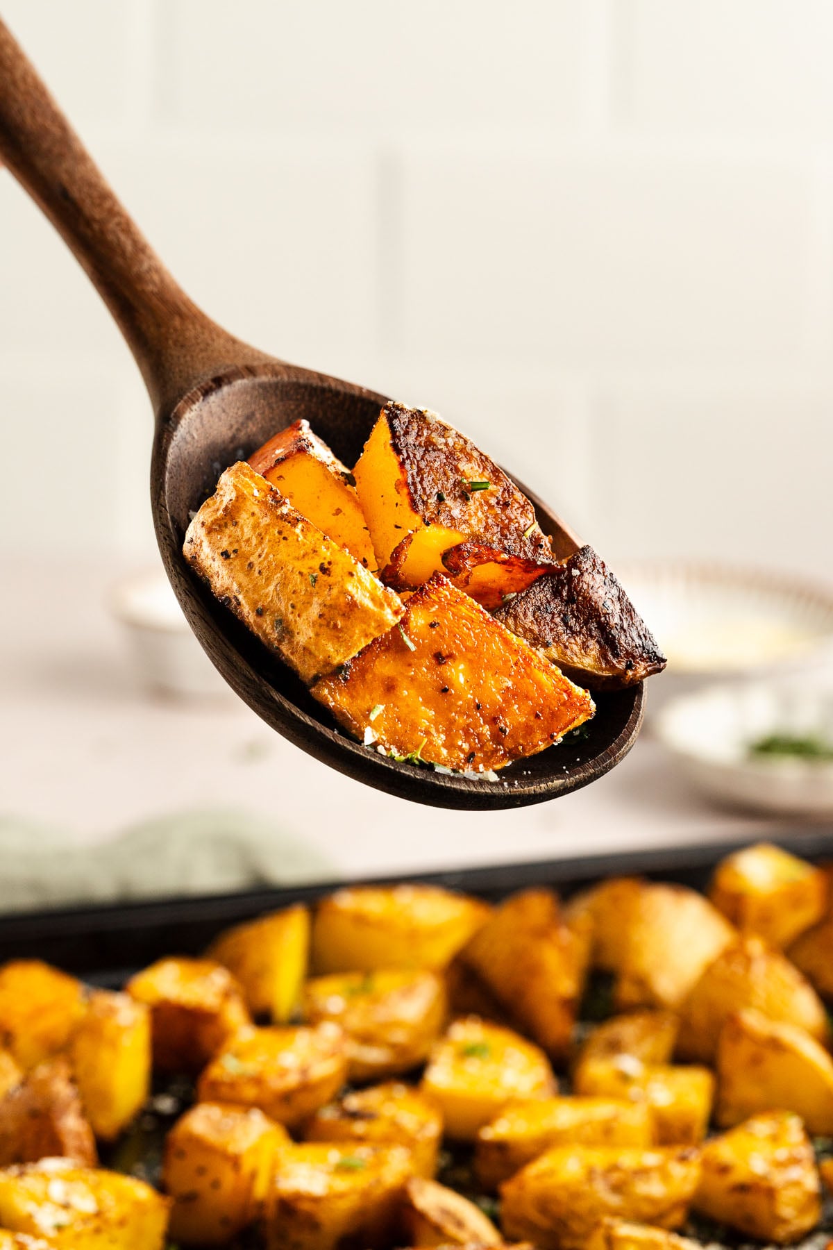 A wooden spoon holds oven crispy oven roasted potato pieces above a tray filled with more of them.