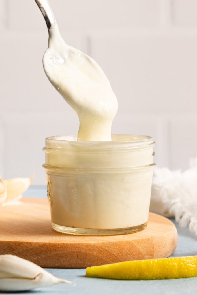 A spoon lifts thick, creamy white sauce from a small glass jar on a wooden board, with a lemon wedge and garlic nearby.