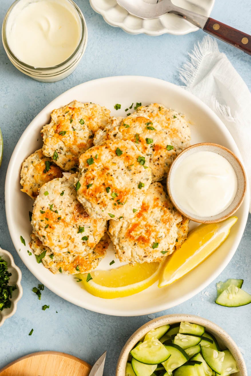 A plate of baked fish cakes garnished with parsley, served with lemon wedges and a small bowl of dipping sauce; cucumber salad and sauce are nearby.