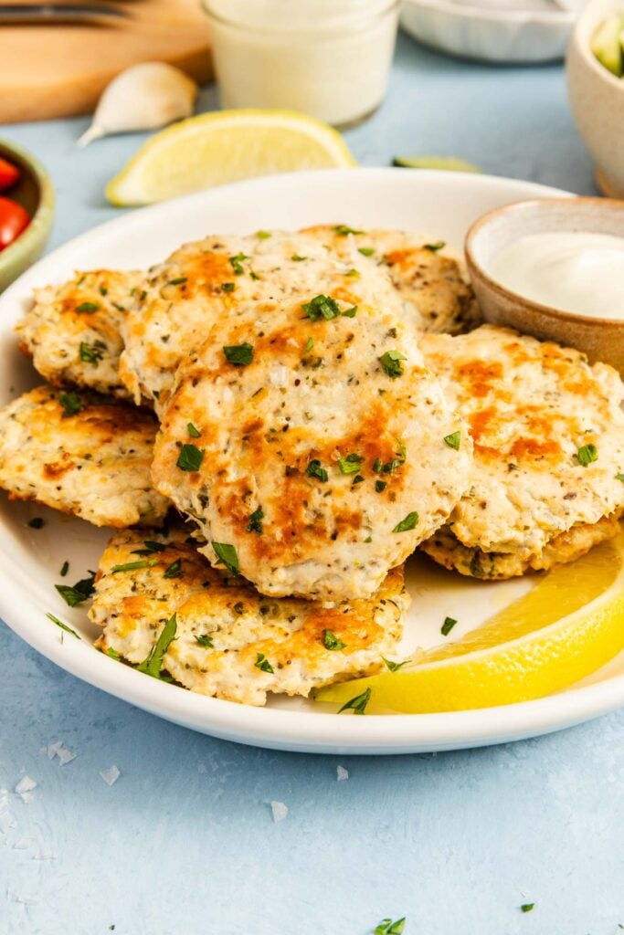 A plate of golden-brown chicken patties garnished with chopped herbs, served with lemon wedges and a small bowl of white dipping sauce.