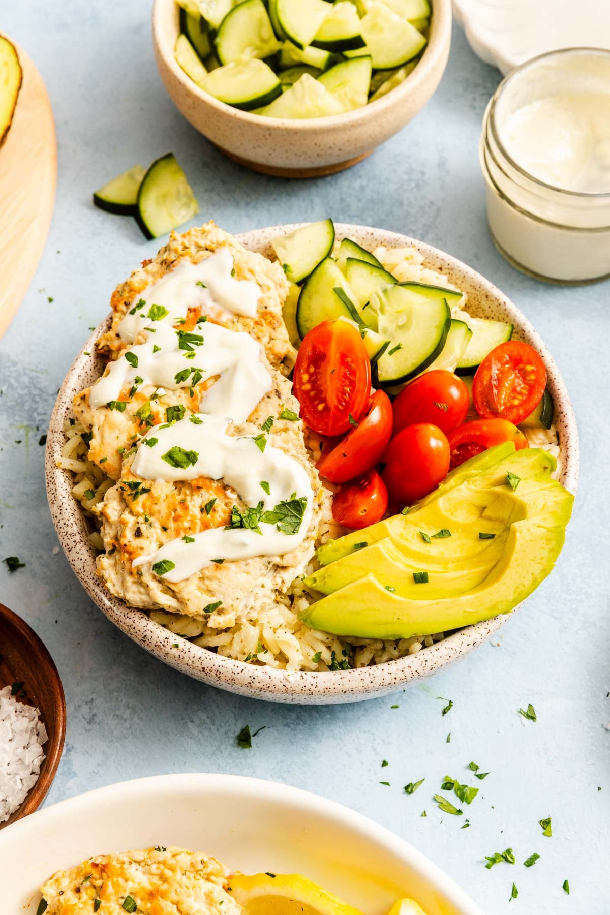 A bowl with grilled chicken patties, sliced avocado, cherry tomatoes, cucumber slices, fluffy white rice, and a drizzle of white sauce, garnished with chopped herbs. Other bowls with cucumbers and sauce nearby.