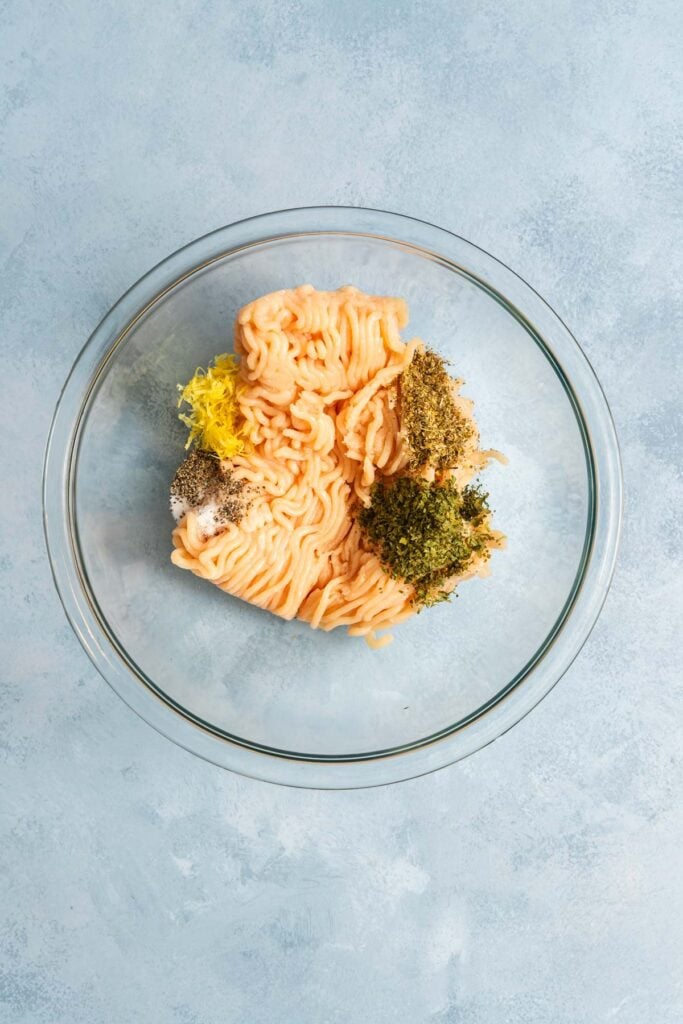 Ground meat with various herbs and spices is placed in a clear glass bowl on a light blue textured surface.