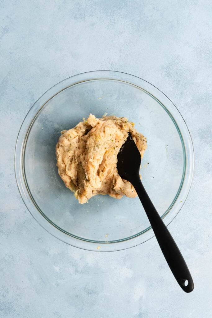 A glass bowl containing a mixture of dough with herbs, being stirred with a black spatula, on a light blue surface.