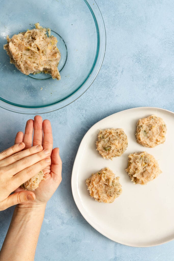Hands shaping raw chicken mixture into patties; four formed patties sit on a white plate, with remaining mixture in a glass bowl on a blue surface.
