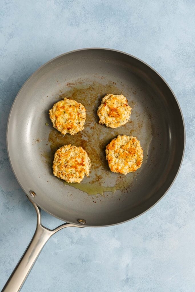 Four golden-brown patties are cooking in a nonstick frying pan on a light blue surface.