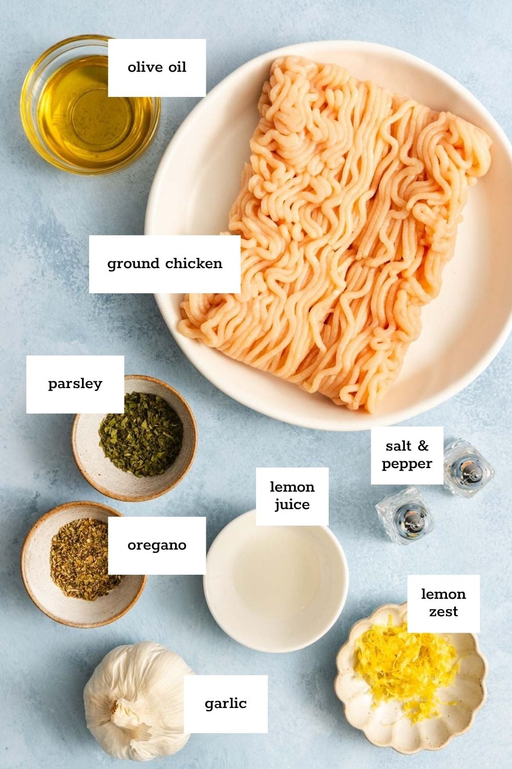 Ingredients for a recipe are arranged on a blue surface, including ground chicken, olive oil, parsley, oregano, lemon juice, lemon zest, garlic, salt, and pepper.
