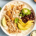 A bowl of white rice topped with shredded meat, black beans, sliced avocado, jalapeño, cilantro, crumbled cheese, and a lime wedge.