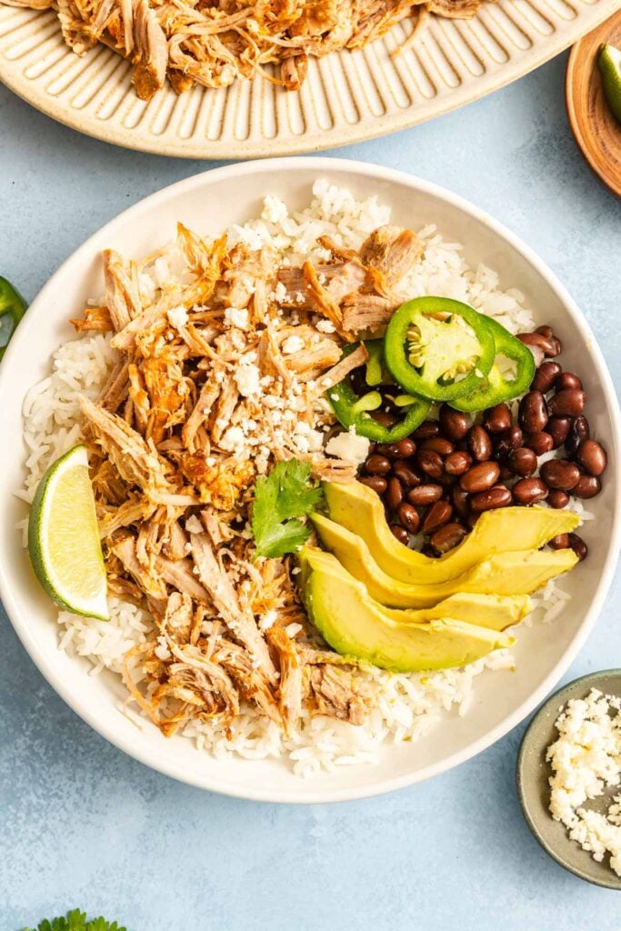 A bowl of white rice topped with shredded meat, black beans, sliced avocado, jalapeño, cilantro, crumbled cheese, and a lime wedge.