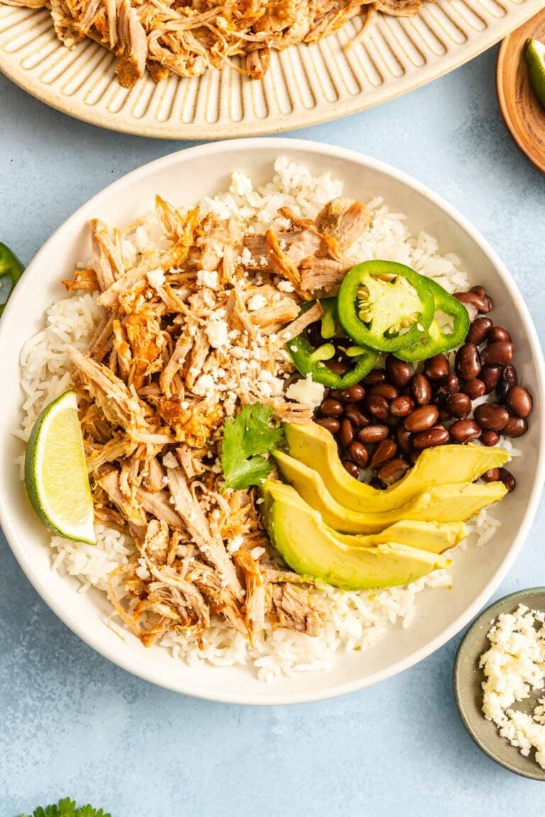 A bowl of white rice topped with shredded meat, black beans, sliced avocado, jalapeño, cilantro, crumbled cheese, and a lime wedge.
