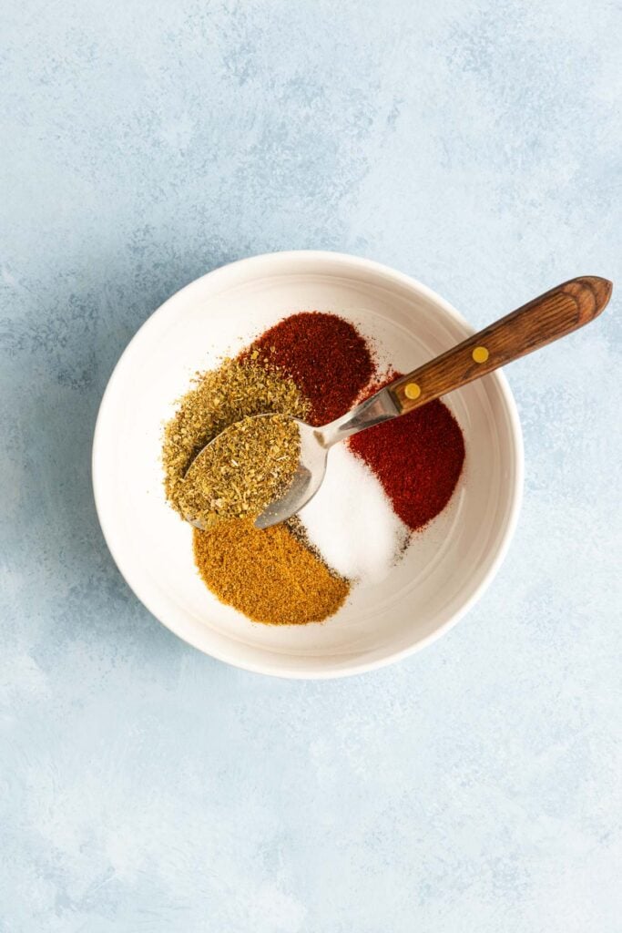 A white bowl containing four piles of spices—dried oregano, chili powder, salt, and cumin—with a metal spoon resting inside.