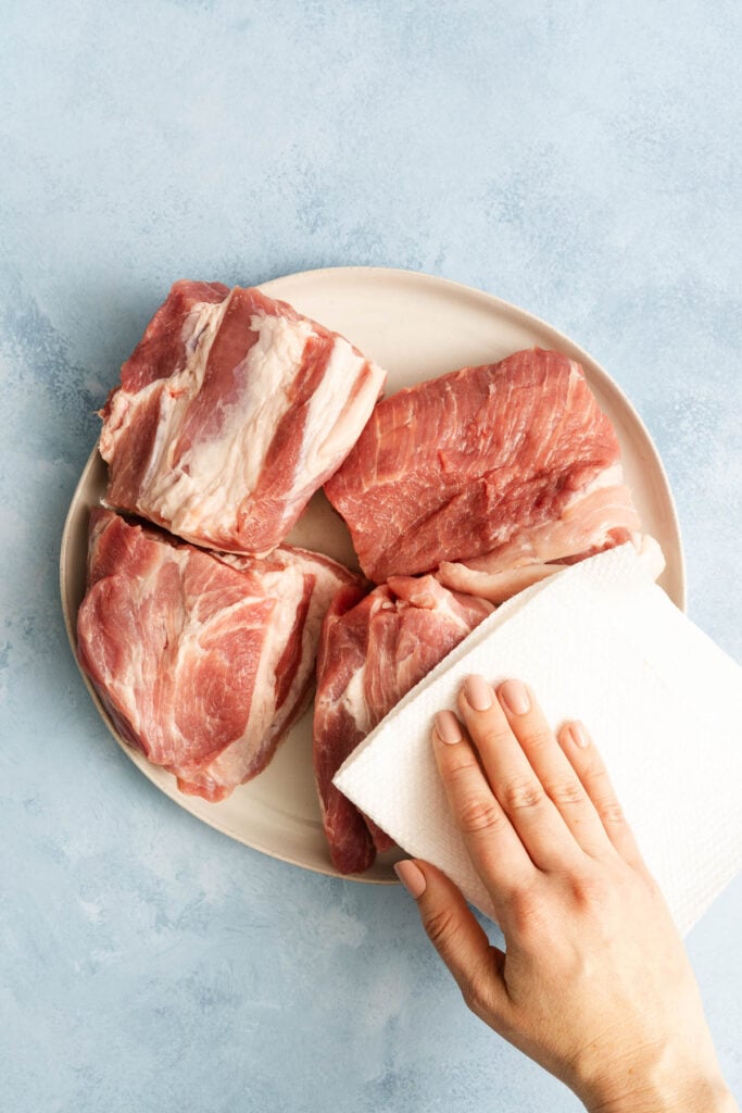 A hand uses a paper towel to pat dry four raw pieces of pork on a white plate against a light blue background.