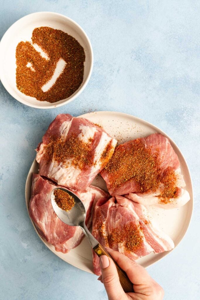 A hand sprinkles seasoning onto four raw pork pieces on a white plate, with a bowl of spice mix beside them on a light blue surface.