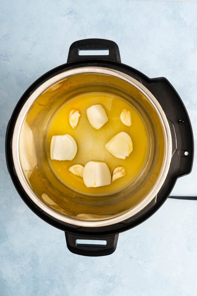 An overhead view of an Instant Pot containing melted butter, onion chunks, and garlic cloves on a light blue surface.