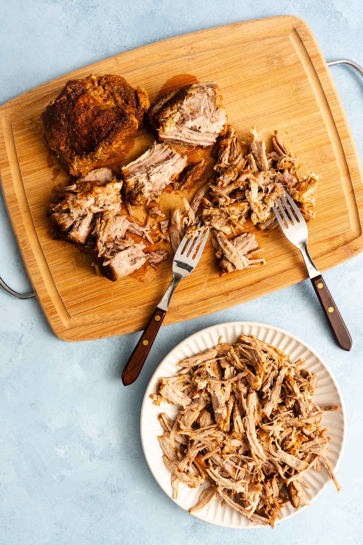Shredded pork being pulled apart with two forks on a wooden cutting board, with more shredded pork served on a white plate below.