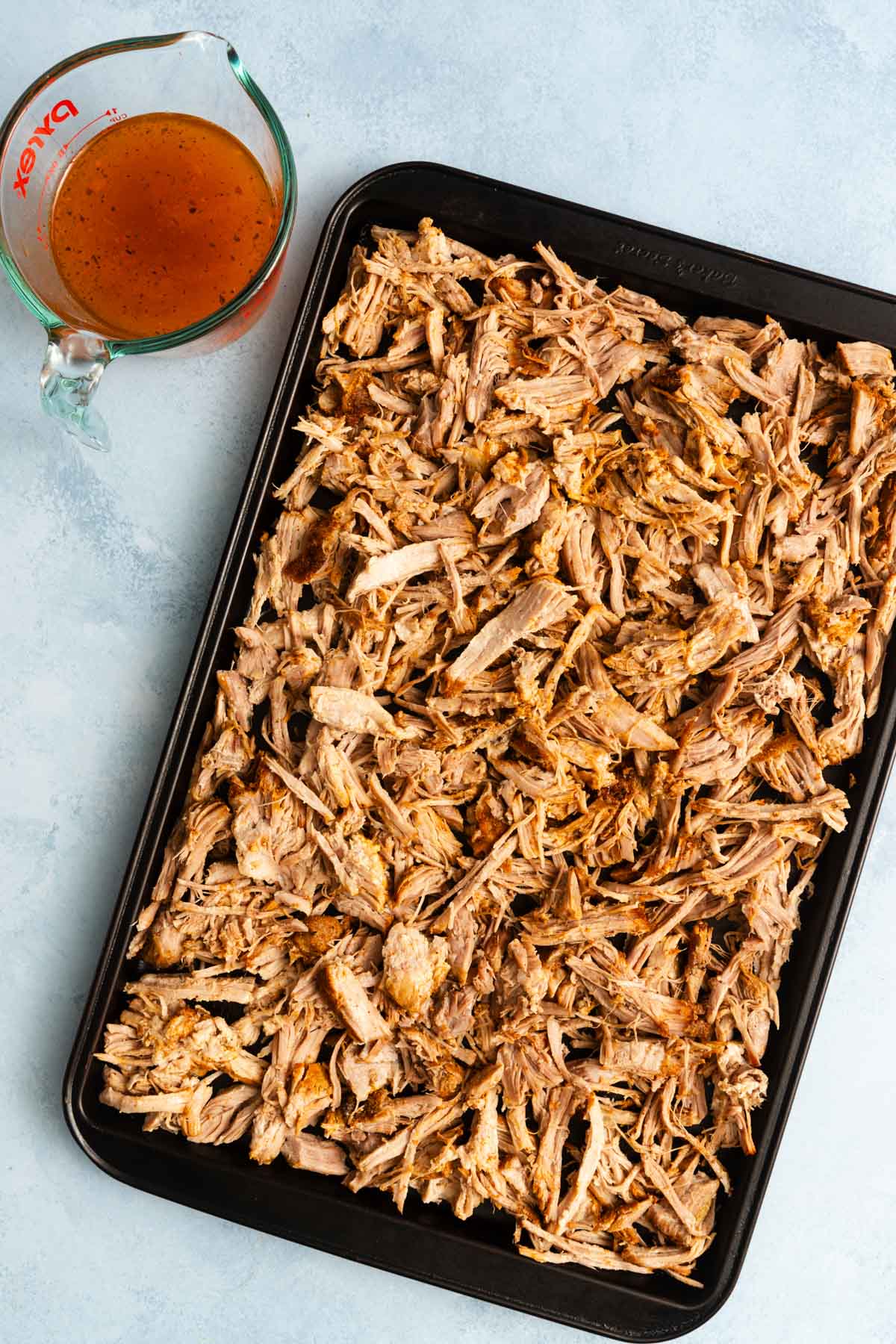 A baking sheet filled with Mexican pulled pork sits next to a glass measuring cup containing brown liquid.