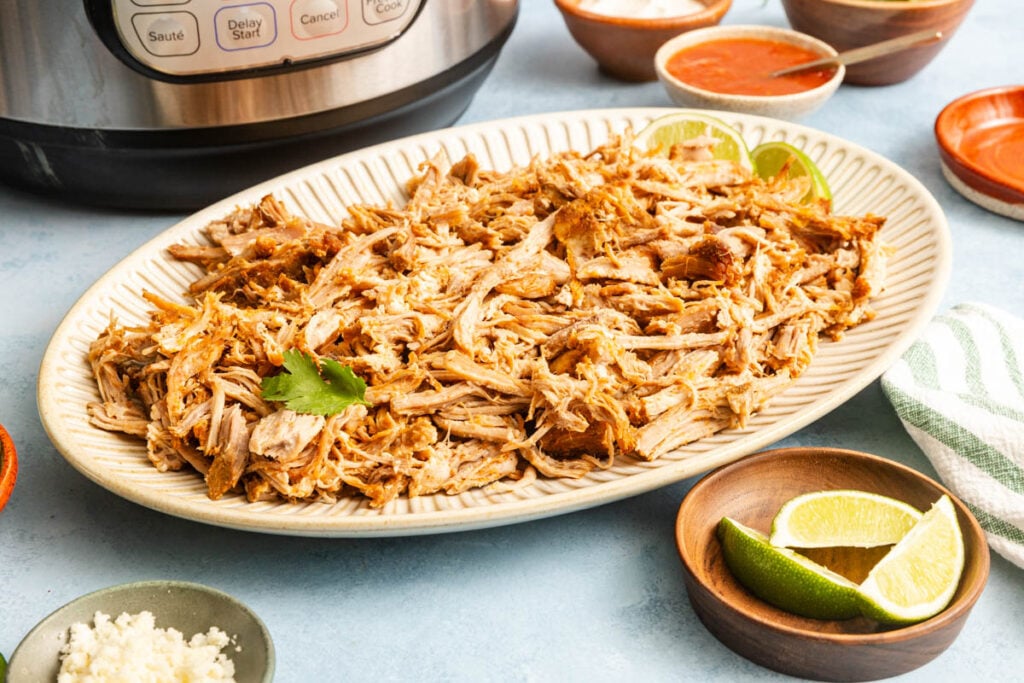 A platter of Instant Pot carnitas garnished with lime wedges and cilantro, surrounded by bowls of sauce, cheese, and limes, with a pressure cooker in the background.