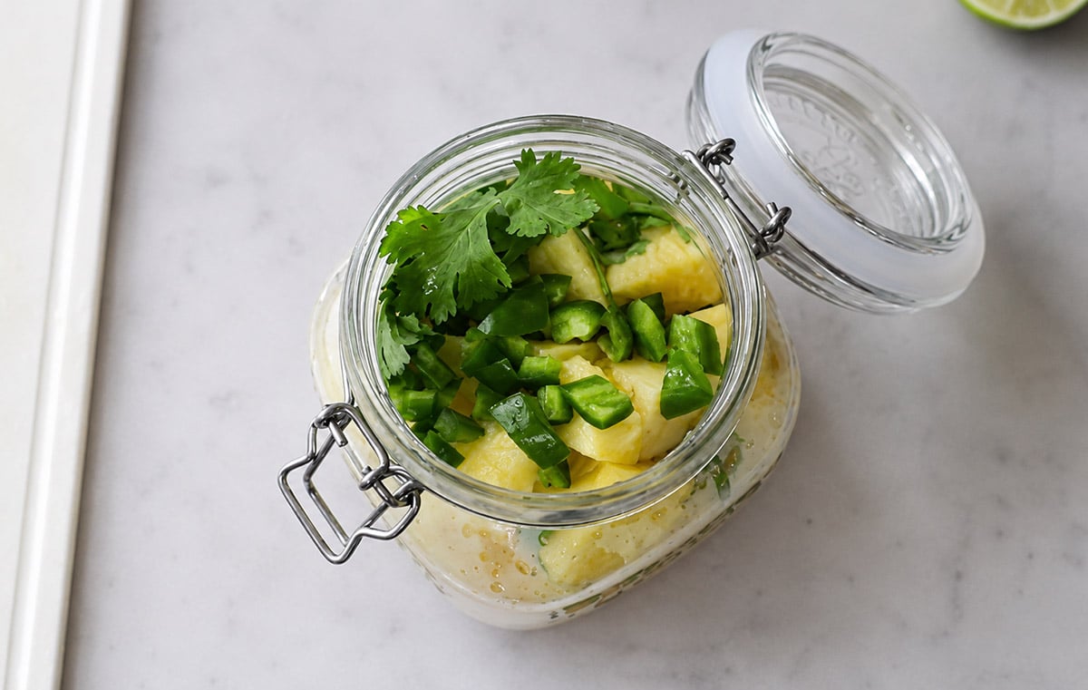 An open glass jar filled with chopped pineapple, green chili peppers, and cilantro sits on a marble surface.
