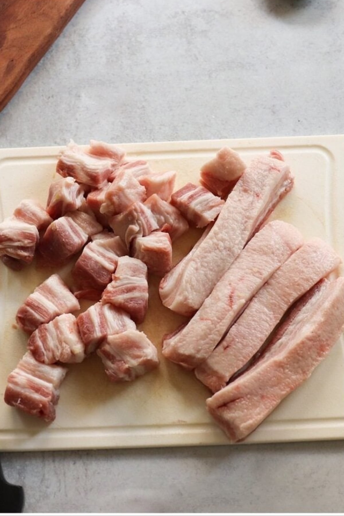 Raw pork belly cut into strips and cubes on a white cutting board, placed on a light-colored surface.