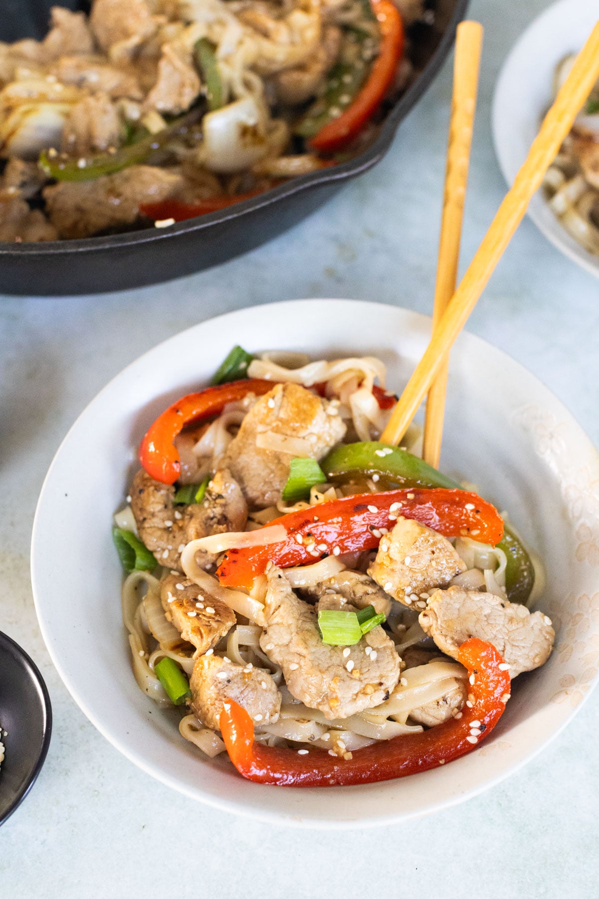 A bowl of stir-fried noodles with sliced pork, red bell peppers, green onions, and sesame seeds, served with chopsticks.