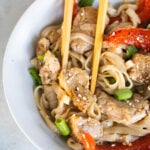 A bowl of noodle stir-fry with pieces of pork tenderloin, red bell pepper, green onions, and sesame seeds, with chopsticks picking up a piece of meat.